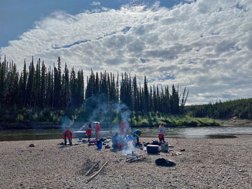 A group of Nę K’ǝ́dı́ Ke Land Guardians standing along the shores of the Dehcho (Mackenzie River) around a fire warming up after practicing whitewater rescue.
